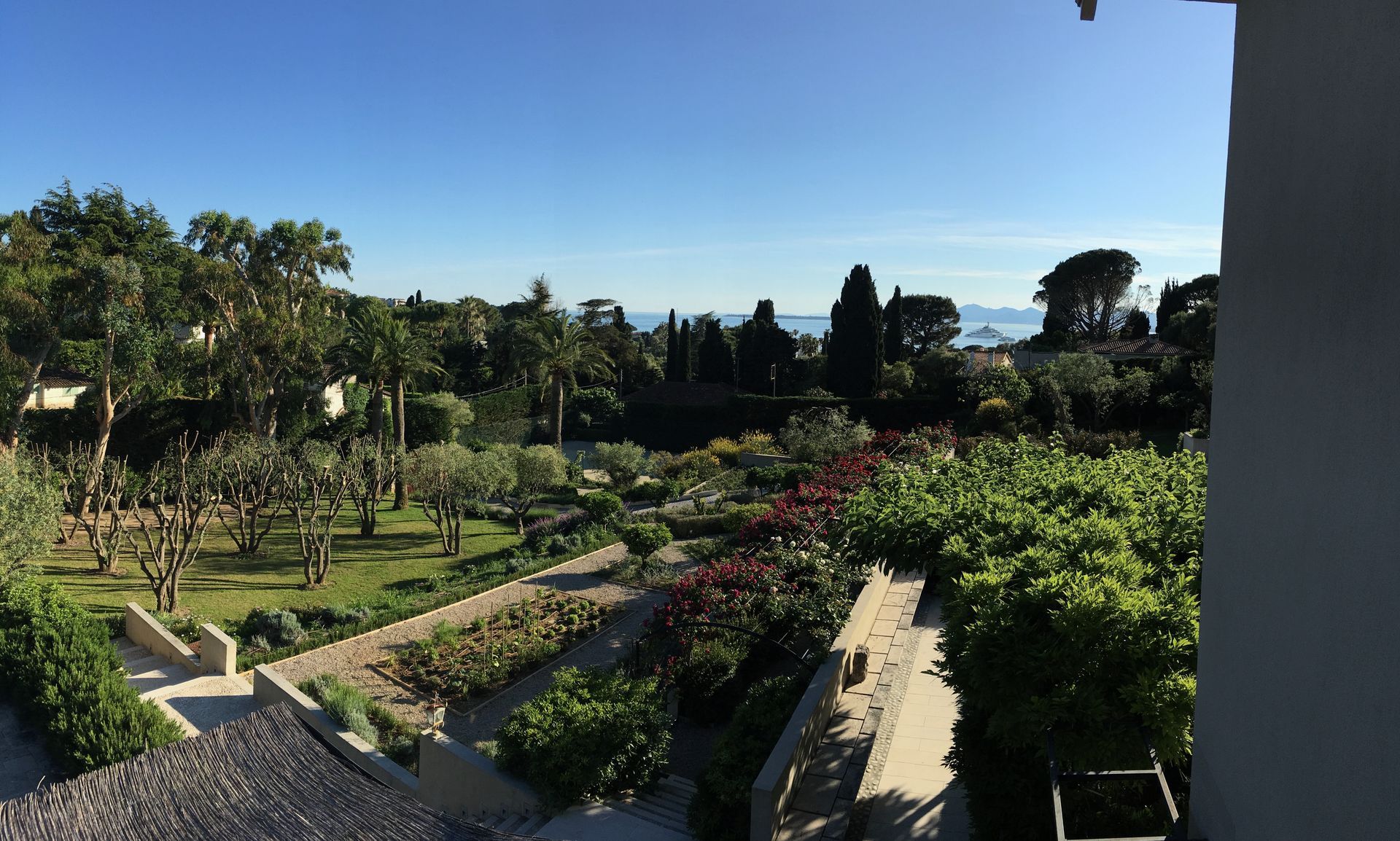 Vue d'un jardin avec des marches, des arbres et l'océan au loin sous un ciel bleu limpide.