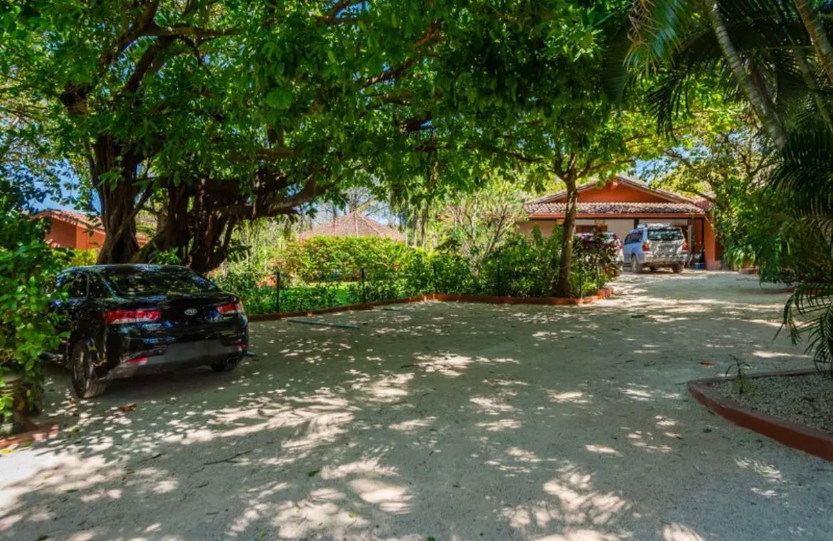 Voiture garée dans une allée de gravier sous des arbres, vue sur une maison.