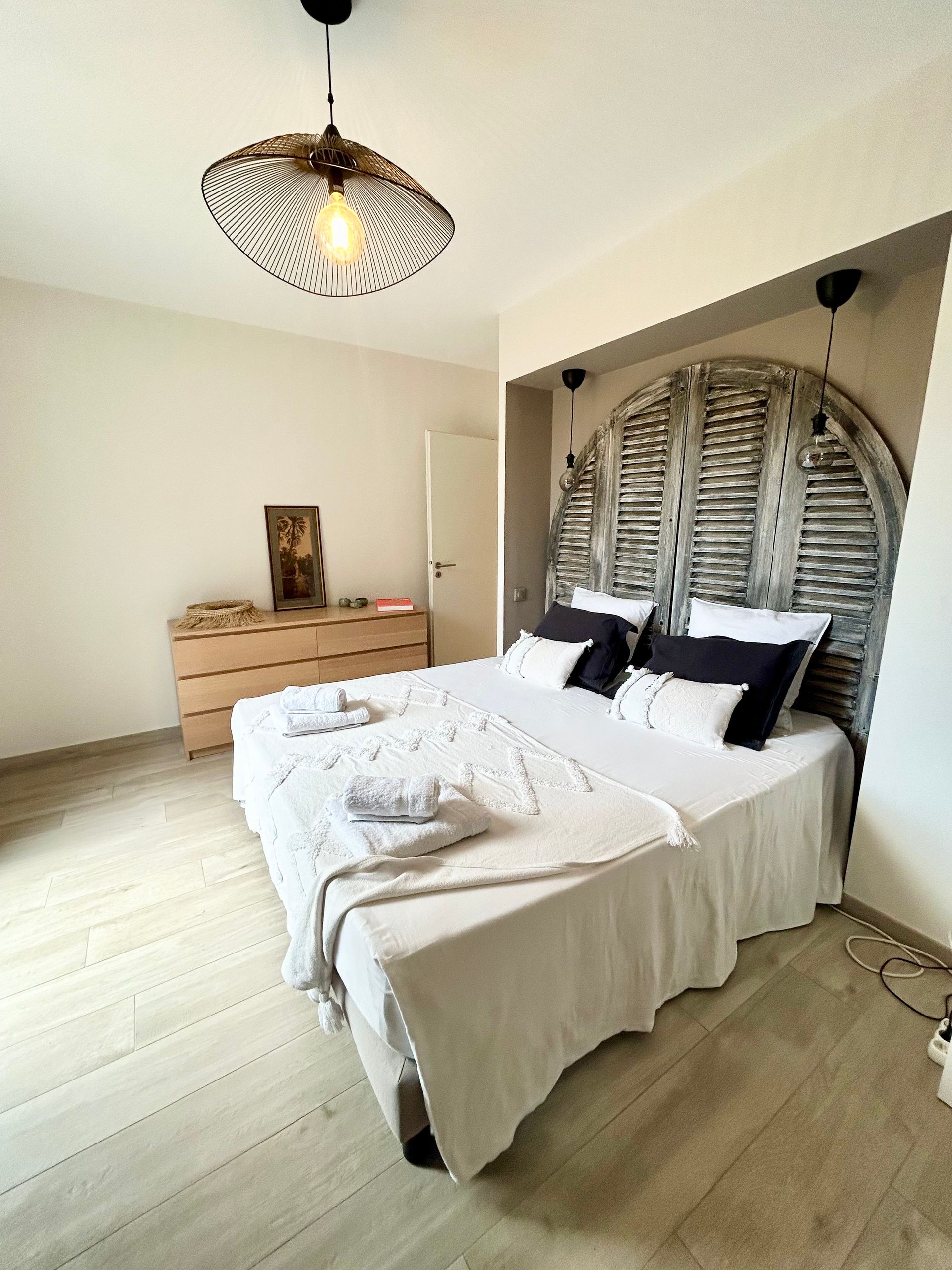 Chambre avec lit, tête de lit, commode et luminaire à motifs. Tons blancs et neutres.