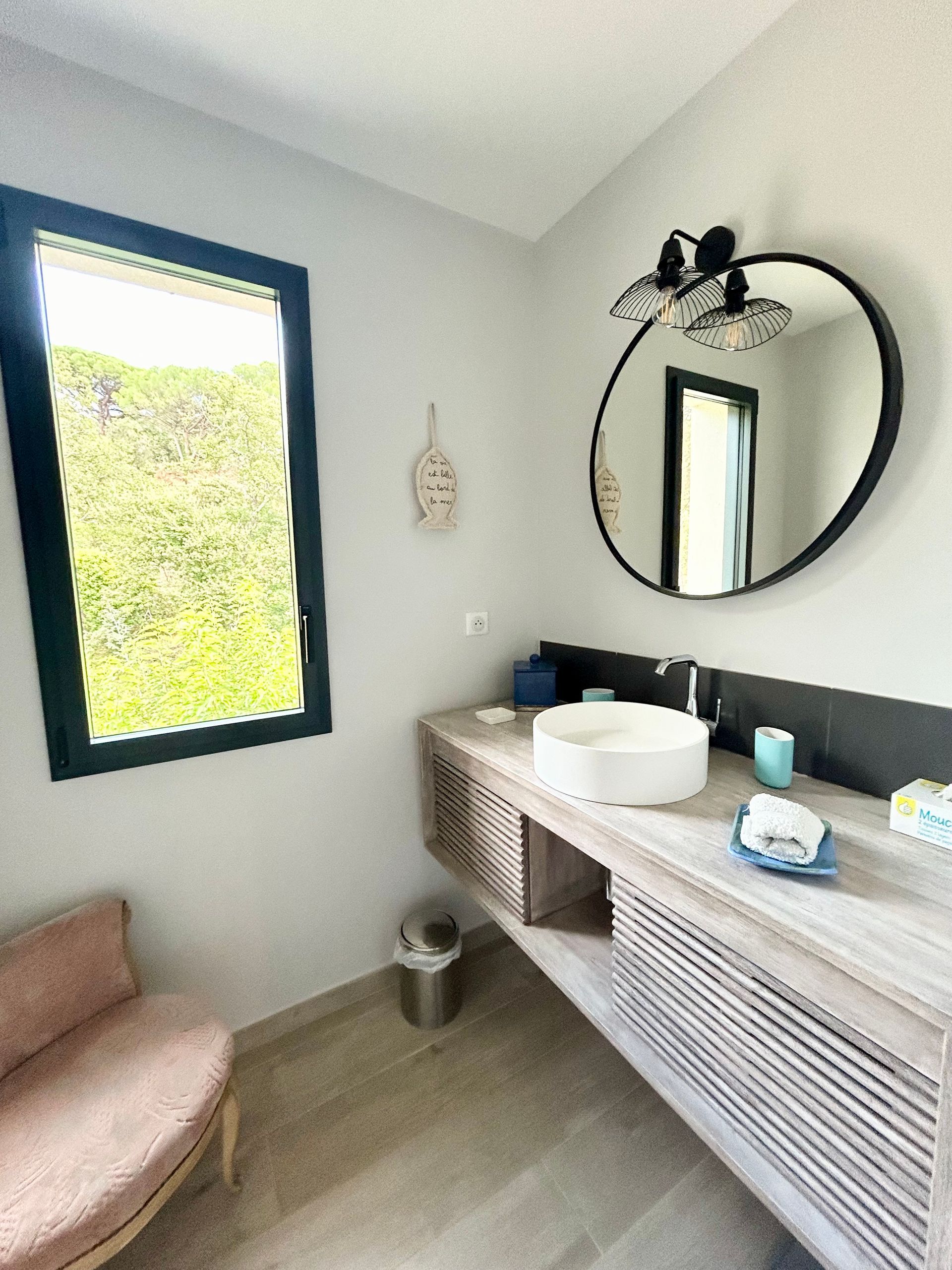 Salle de bains avec fenêtre, miroir rond et meuble-lavabo en bois. Une petite chaise est placée dans un coin.