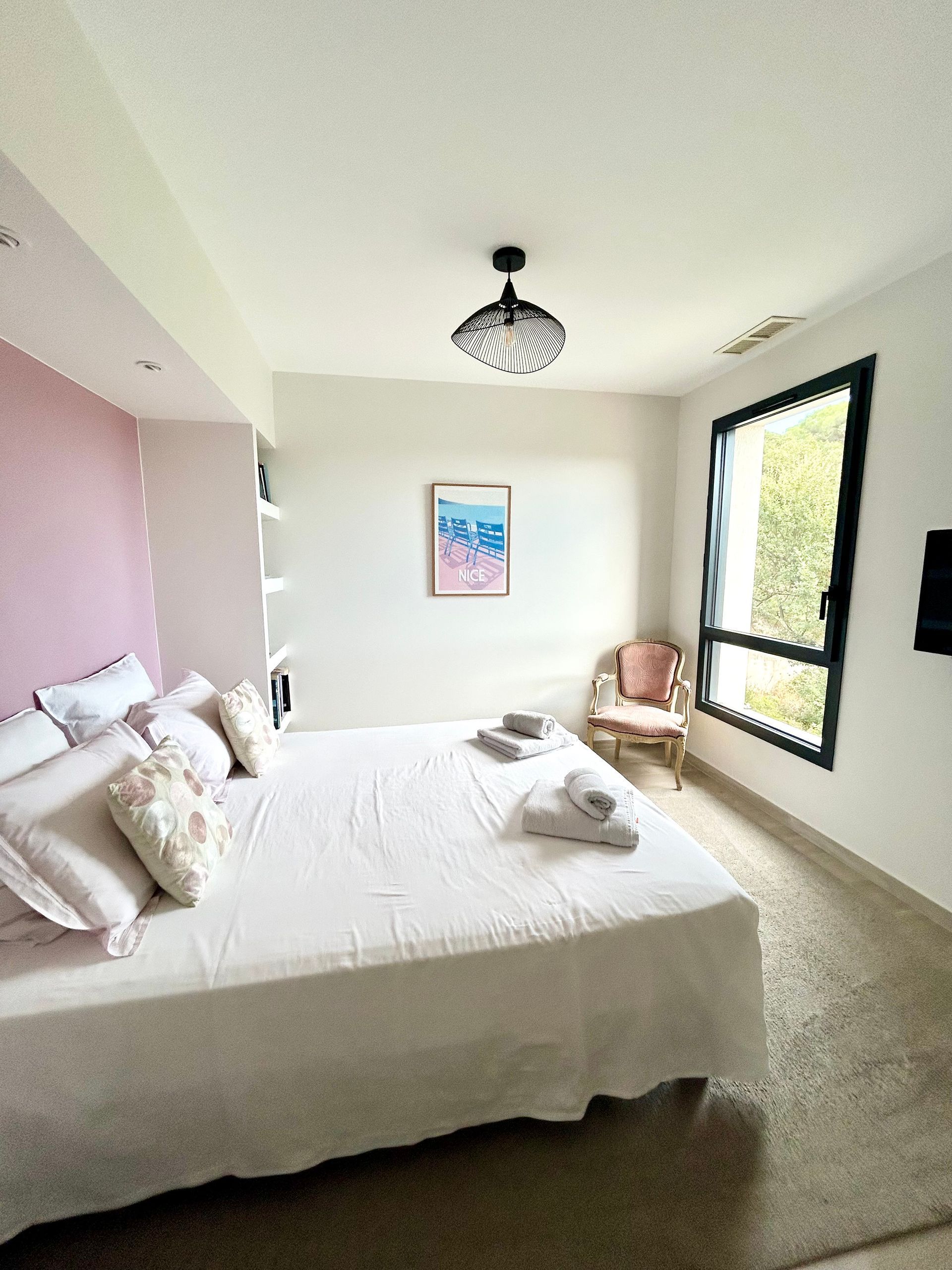 Chambre avec literie blanche, mur rose, cadre de fenêtre noir et chaise rose.
