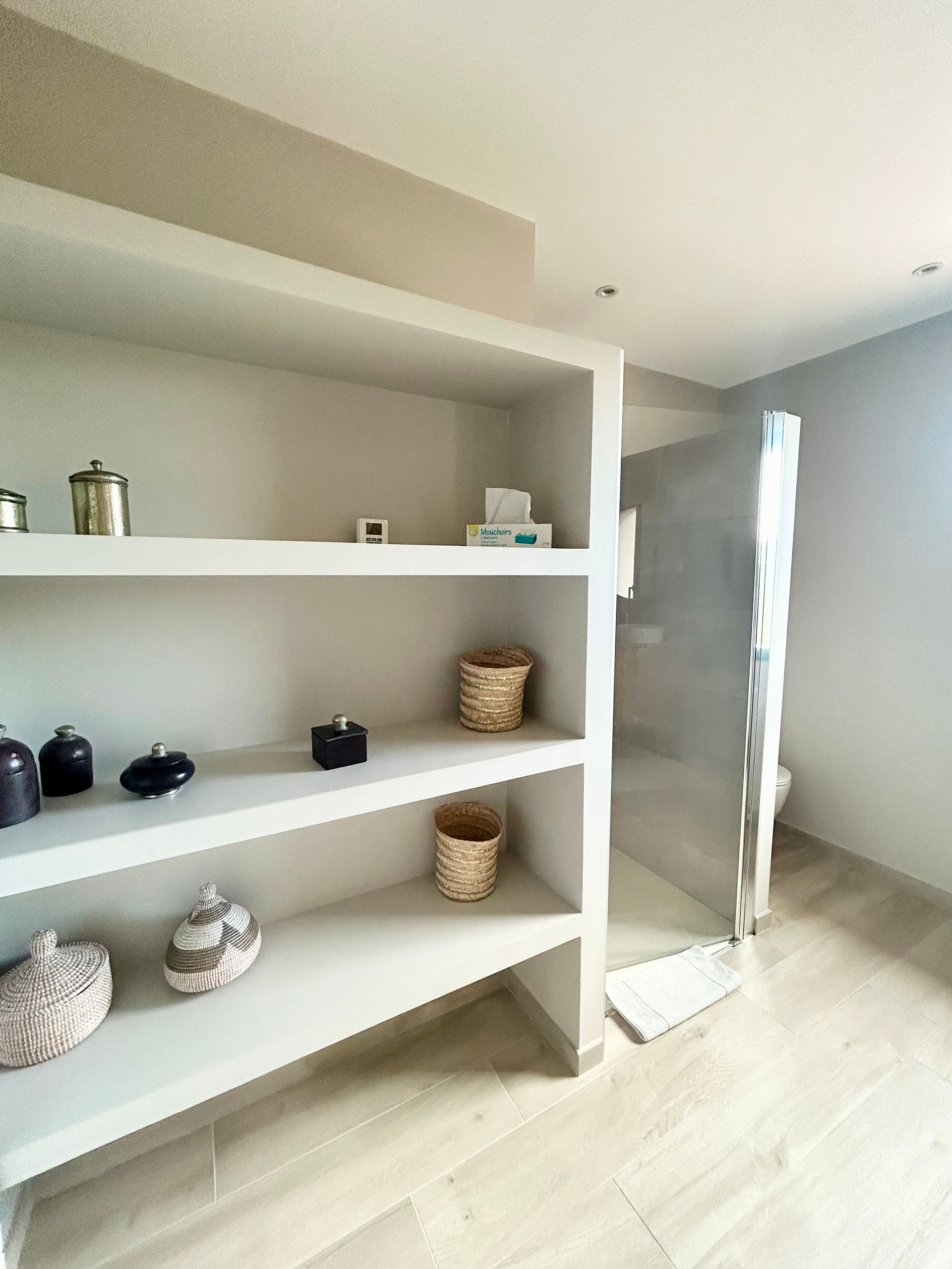 Étagères blanches encastrées avec éléments décoratifs dans une salle de bains aux tons clairs. Sol en bois et espace douche visibles.