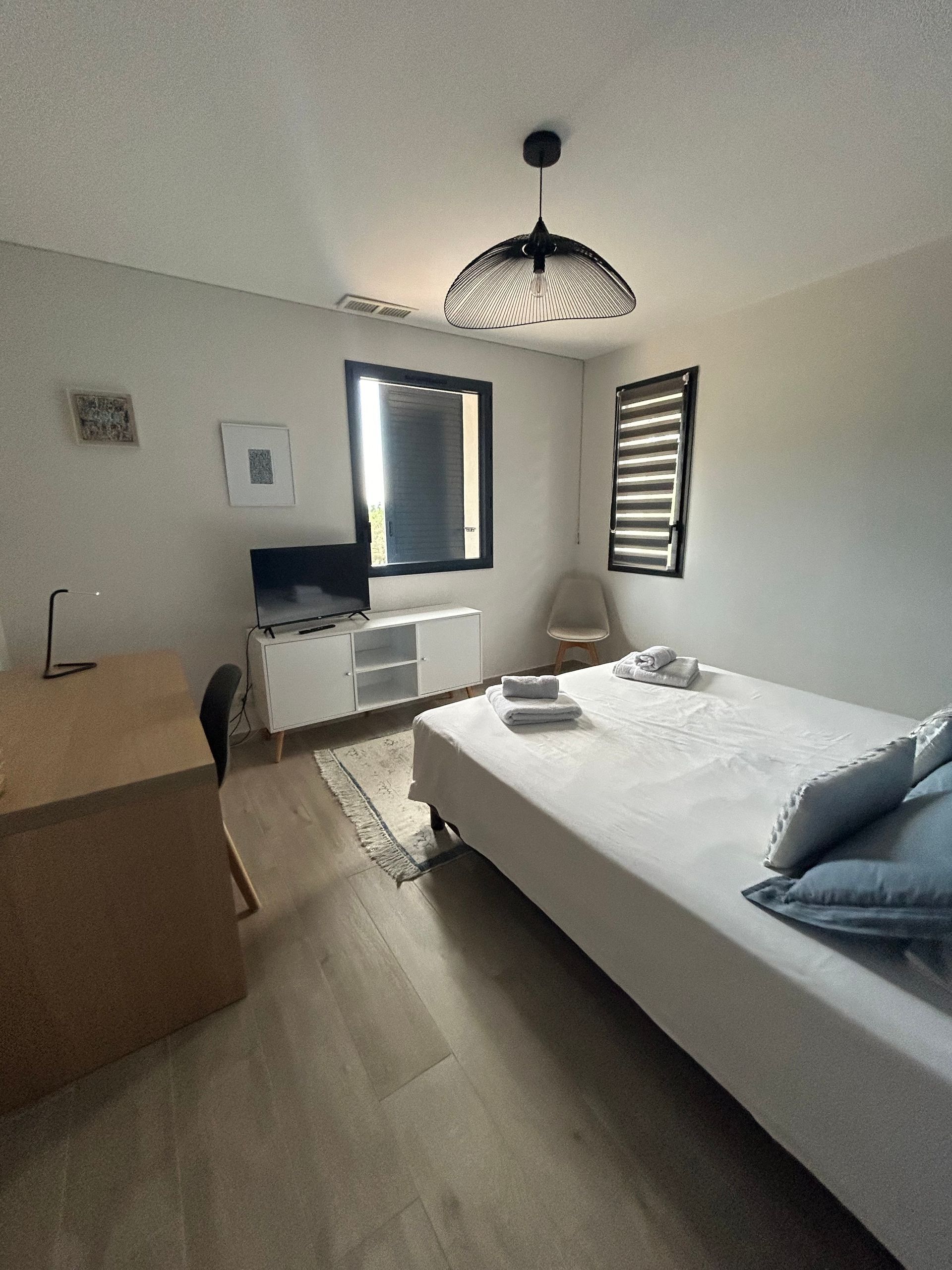 Chambre avec lit, bureau, télévision et fenêtre avec stores.