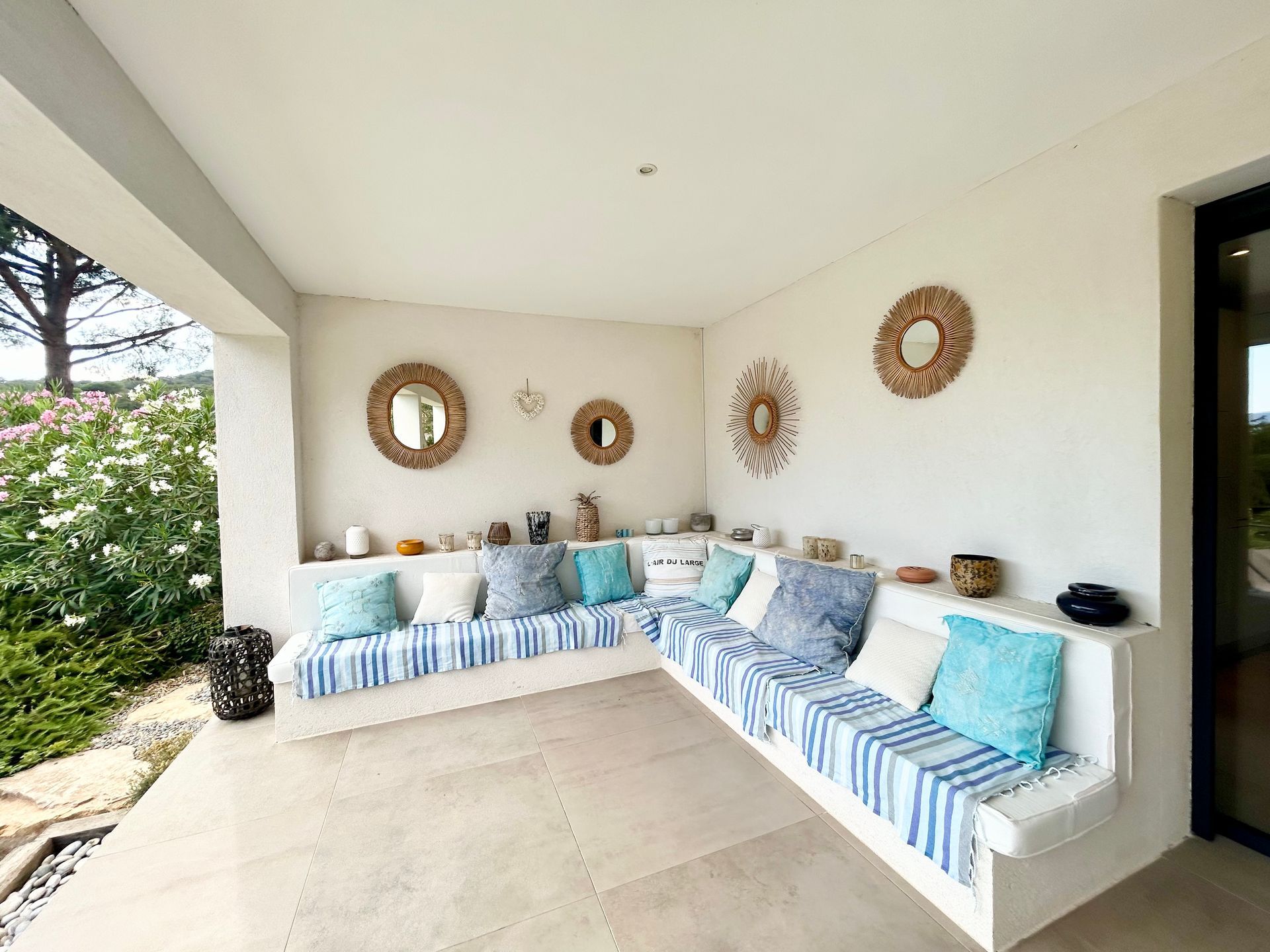 Terrasse extérieure avec banquettes blanches intégrées, coussins à rayures bleues et blanches, miroirs décoratifs et sol carrelé aux tons neutres.