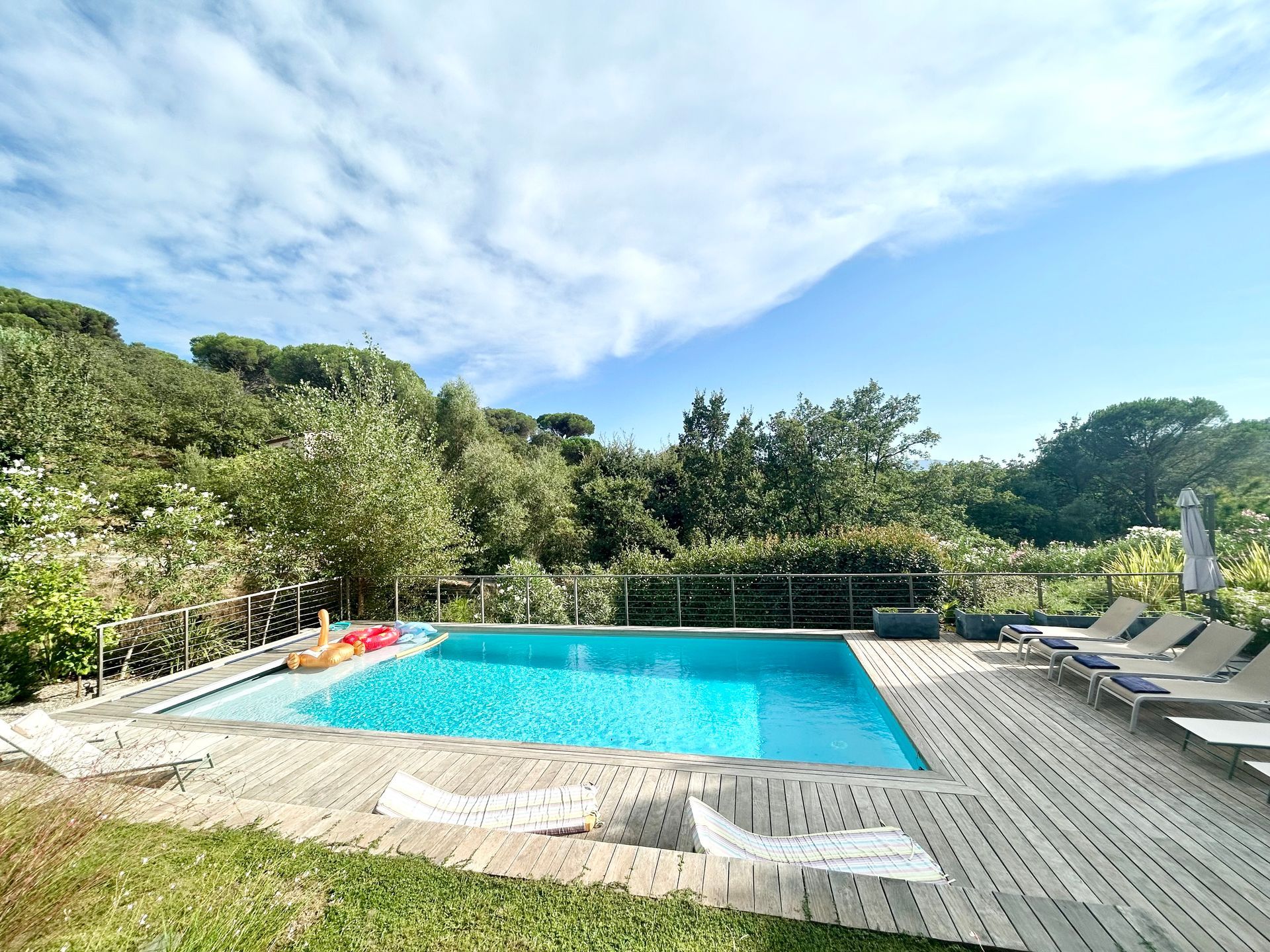 Piscine entourée d'une terrasse en bois, de chaises longues et d'une végétation luxuriante sous un ciel bleu partiellement nuageux.