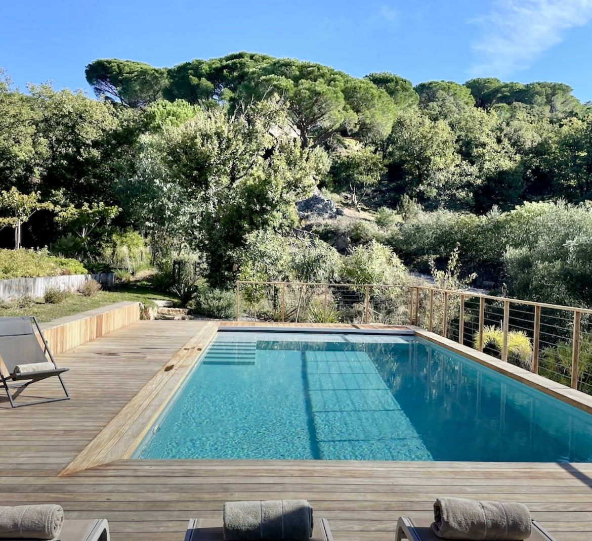 Piscine entourée d'une terrasse en bois et de chaises longues, avec une végétation luxuriante en arrière-plan sous un ciel ensoleillé.
