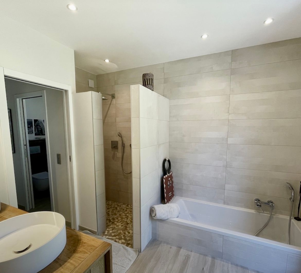 Salle de bains moderne avec douche à l'italienne, baignoire et lavabo suspendu. Murs carrelés gris clair et palette de couleurs neutres.
