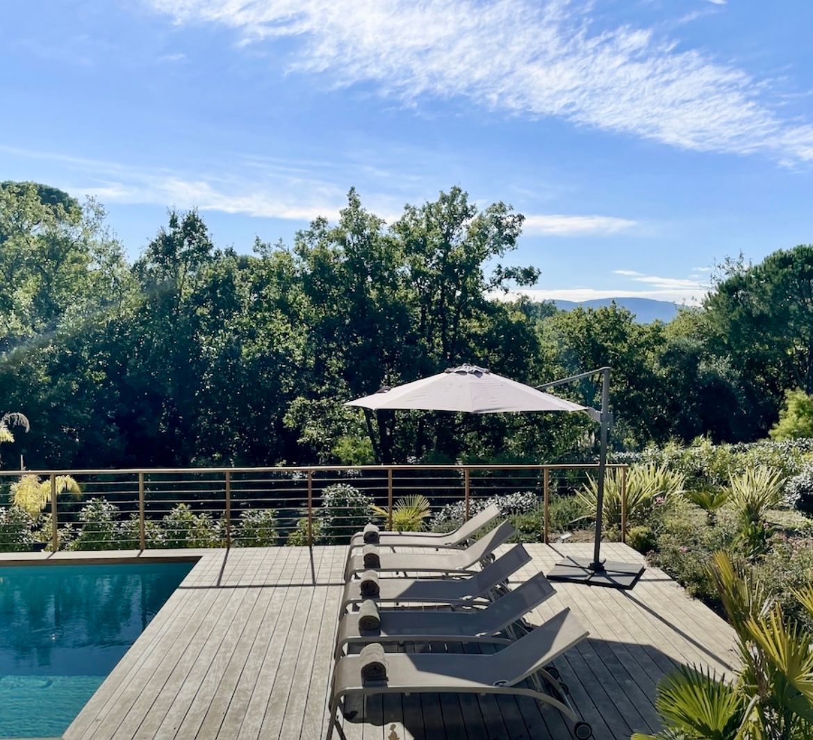 Scène au bord de la piscine avec chaises longues, parasol et vue sur les arbres et le ciel.