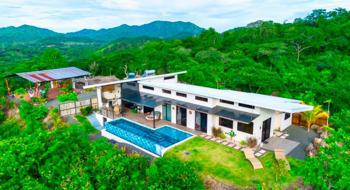 Maison moderne avec piscine et panneaux solaires, entourée d'une végétation luxuriante à flanc de colline.