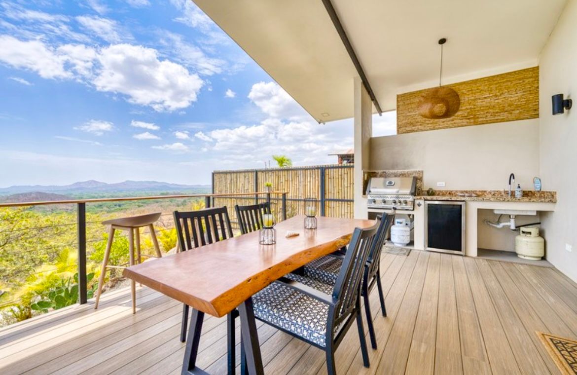 Espace repas extérieur avec barbecue, évier et table en bois donnant sur un paysage pittoresque.
