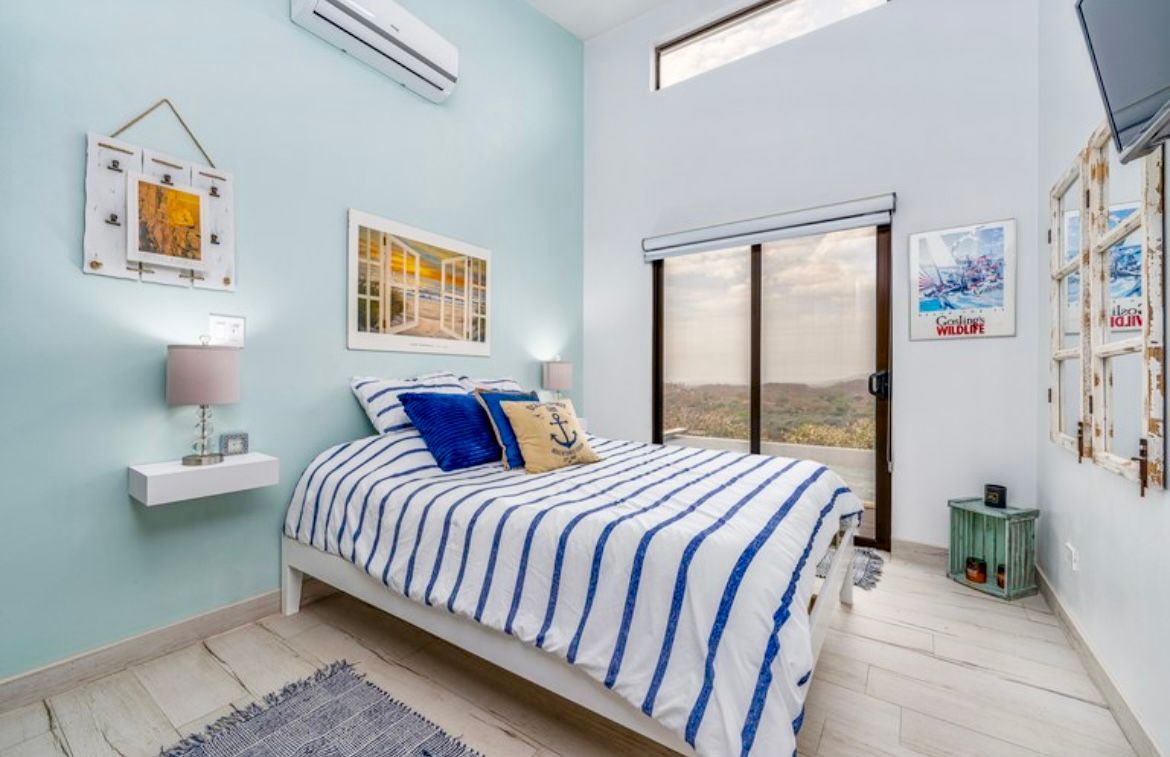Chambre avec linge de lit à rayures bleues et blanches, œuvres d'art et porte-fenêtre coulissante offrant une vue.