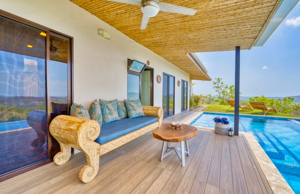 Terrasse extérieure avec piscine. Canapé en osier avec coussins bleus, table en bois et ventilateur de plafond sous un toit en osier.
