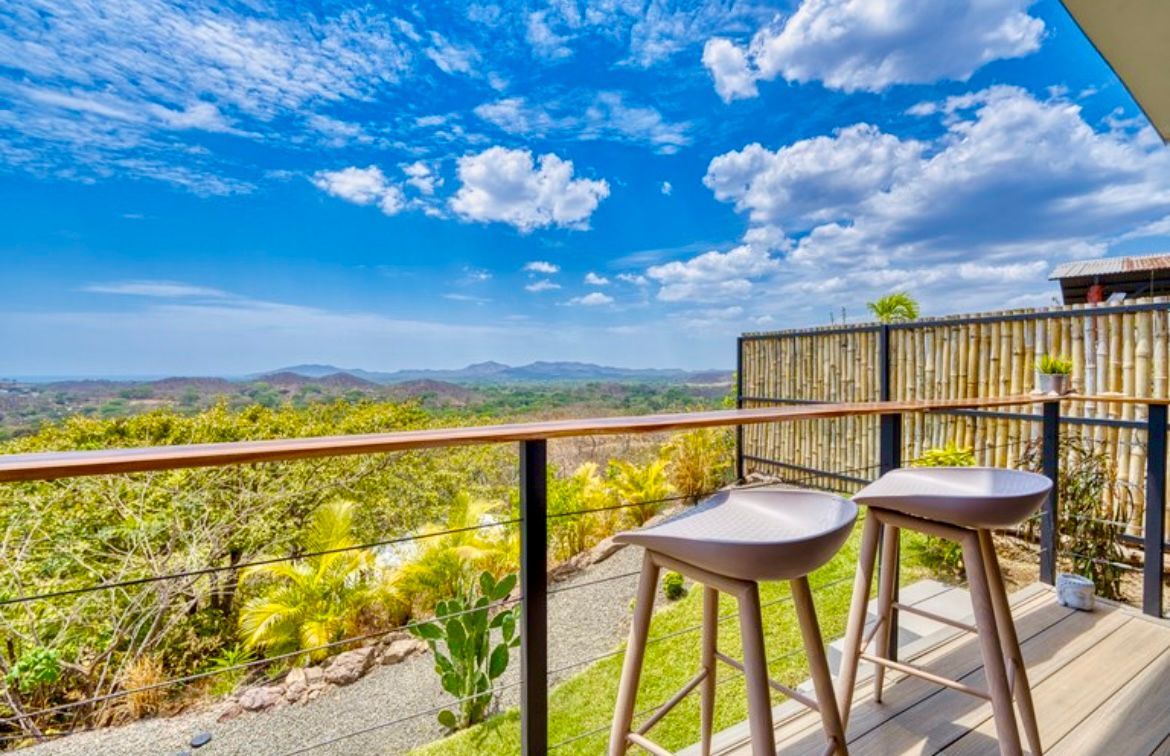 Balcon avec deux tabourets, donnant sur des collines verdoyantes et un ciel bleu vif parsemé de nuages.