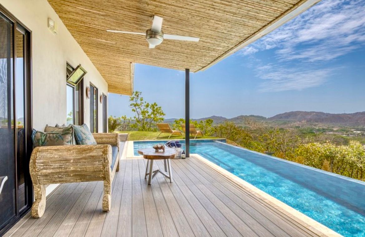 Patio avec piscine donnant sur un paysage ; terrasse en bois, canapé rustique, plafond tissé et ciel bleu.