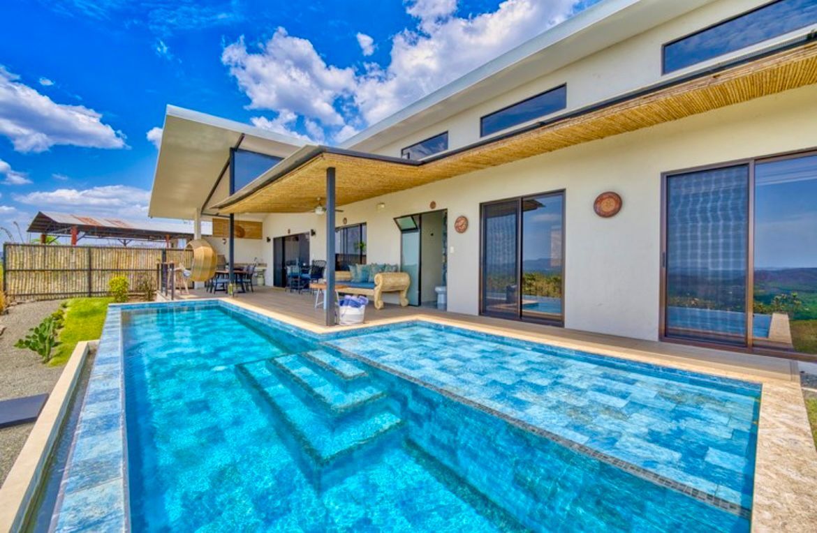 Maison moderne avec une longue piscine carrelée de bleu. Vue extérieure par portes coulissantes ouvertes, ciel dégagé et ensoleillé.