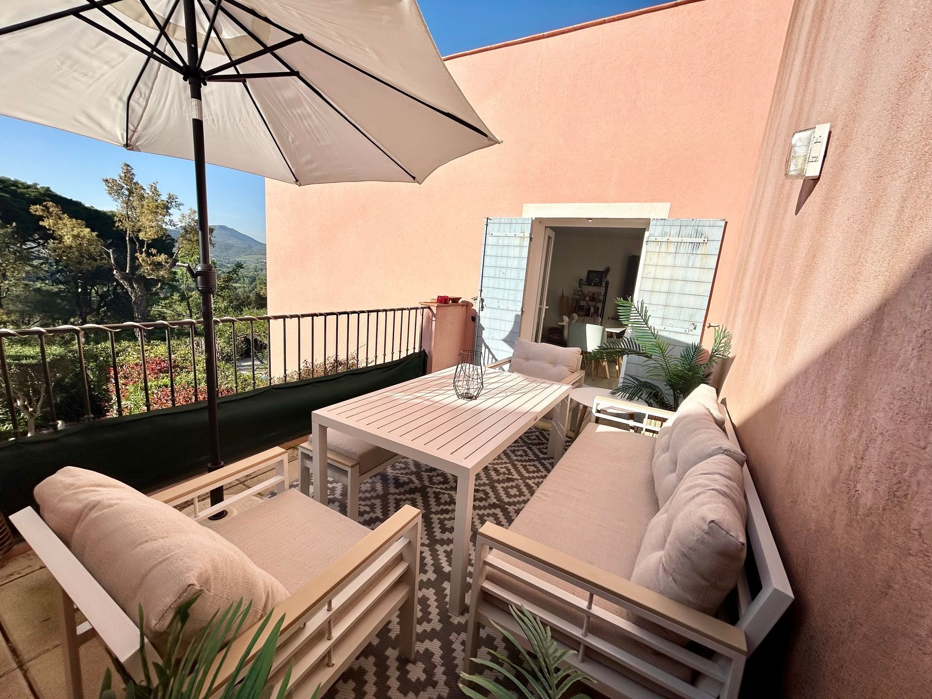 Terrasse aménagée avec table, deux canapés et parasol. Bâtiment rose, porte ouverte, vue sur la montagne.