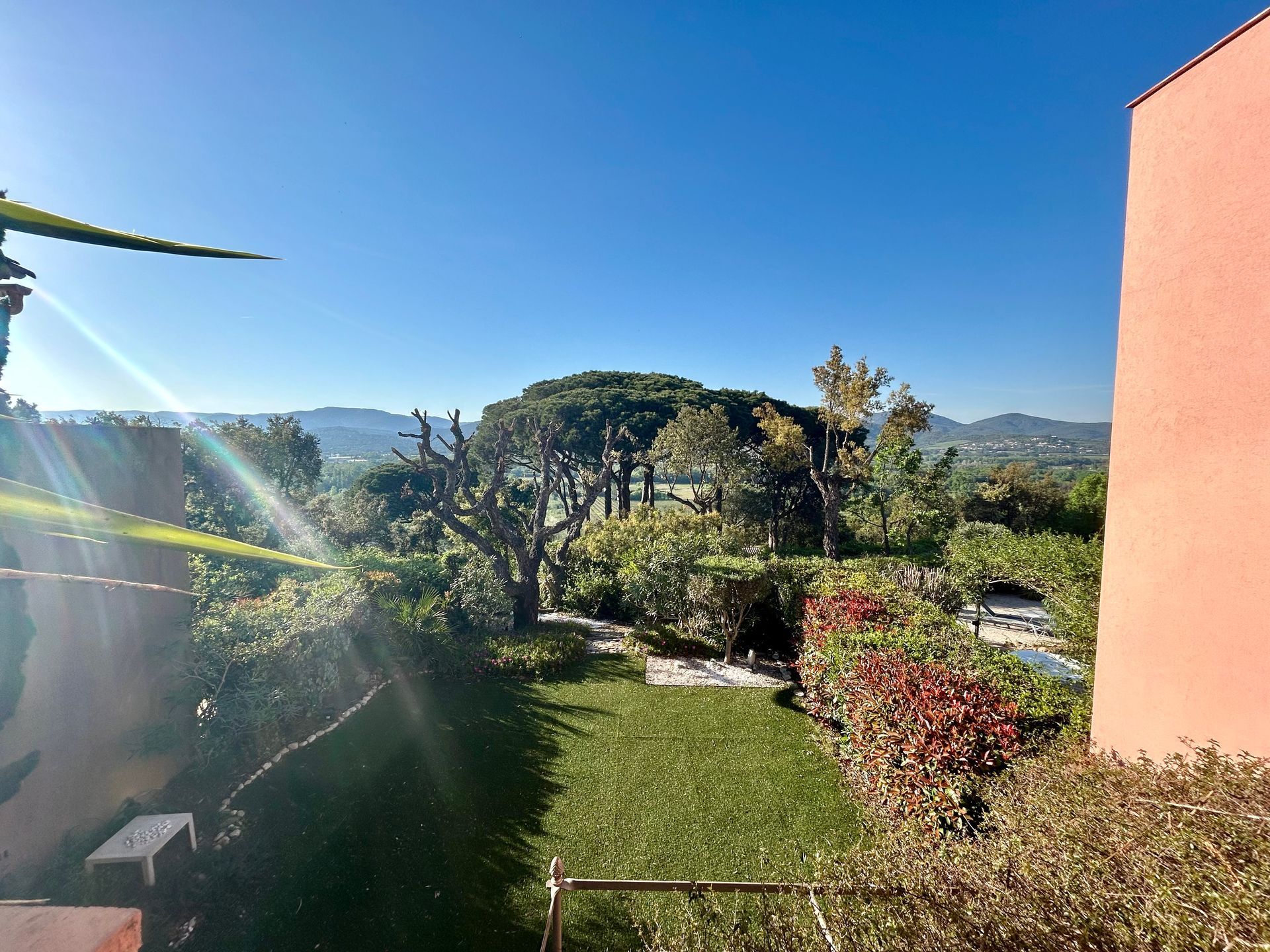 Vue sur un jardin luxuriant, des arbres et des collines au loin sous un ciel d'un bleu éclatant.