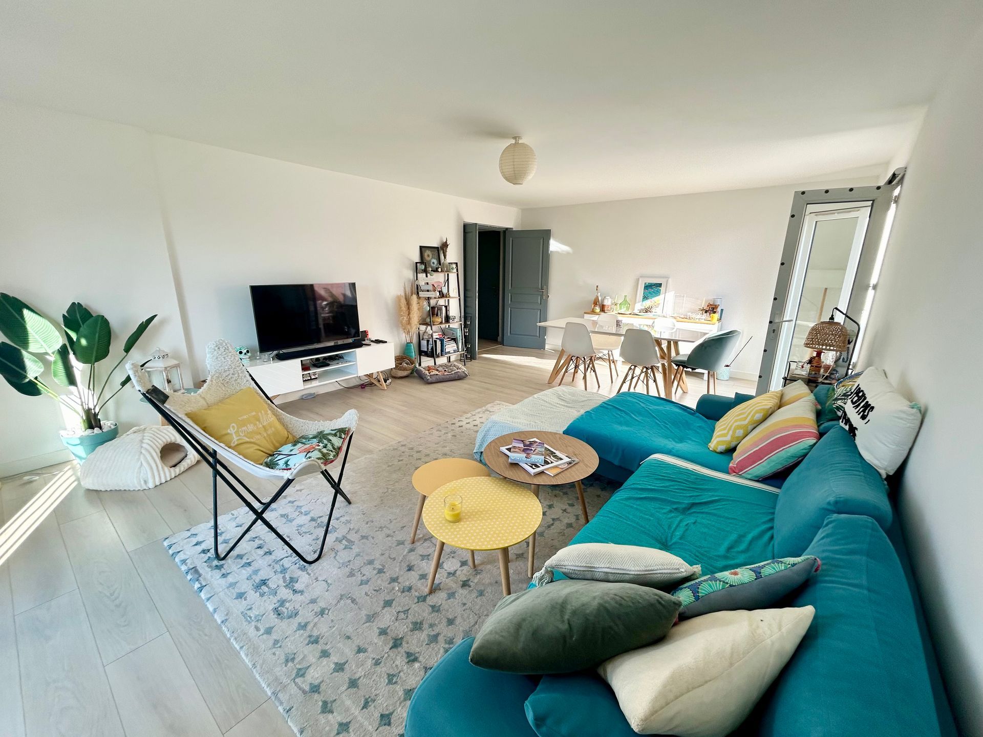 Salon spacieux avec canapé turquoise, télévision et coin repas. Murs aux tons neutres, parquet clair et touches de couleur.
