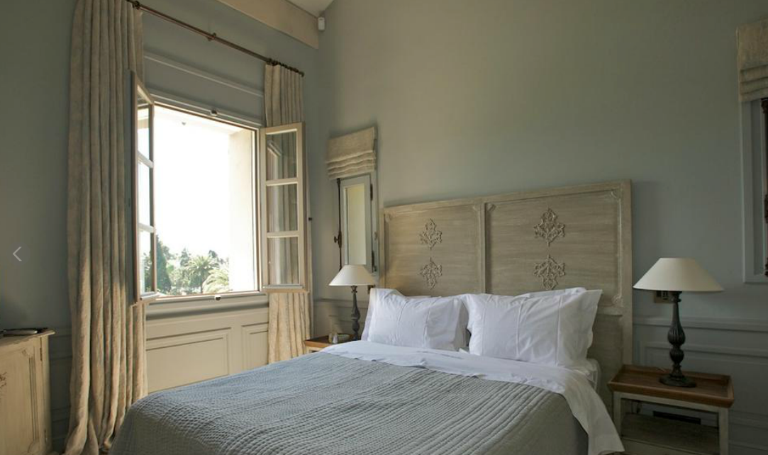 Chambre avec fenêtre ouverte, lit et tables de chevet ; murs bleu clair, rideaux neutres et linge de lit blanc.