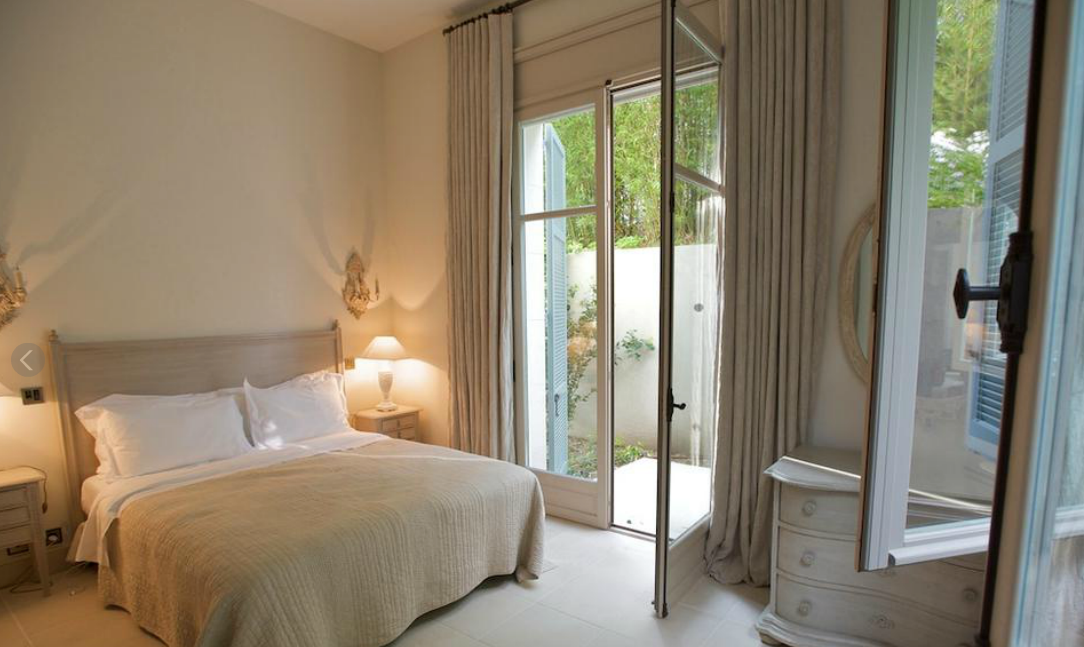 Chambre avec lit, tables de chevet et portes-fenêtres donnant sur un jardin. Tons beiges et blancs.