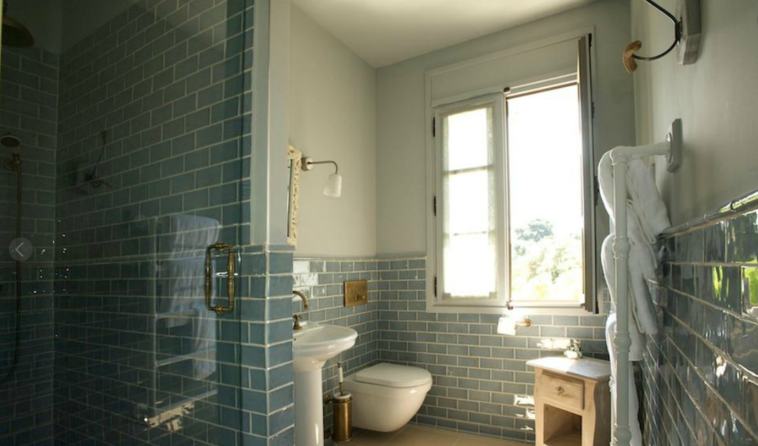 Salle de bains avec murs carrelés bleus, douche en verre, lavabo sur pied et fenêtre laissant entrer la lumière naturelle.