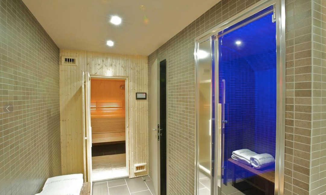 Un couloir menant à un spa avec sauna et hammam éclairé de bleu. Murs carrelés de gris clair.