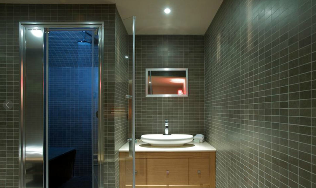 Salle de bains avec murs carrelés gris, douche avec porte en verre, lavabo sur meuble en bois et miroir.