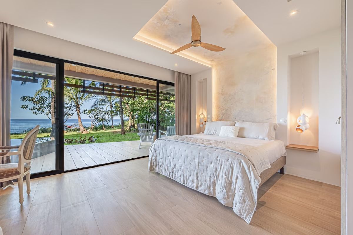 Chambre avec lit king-size, vue sur l'océan, grandes portes vitrées, couleurs neutres et ventilateur de plafond.