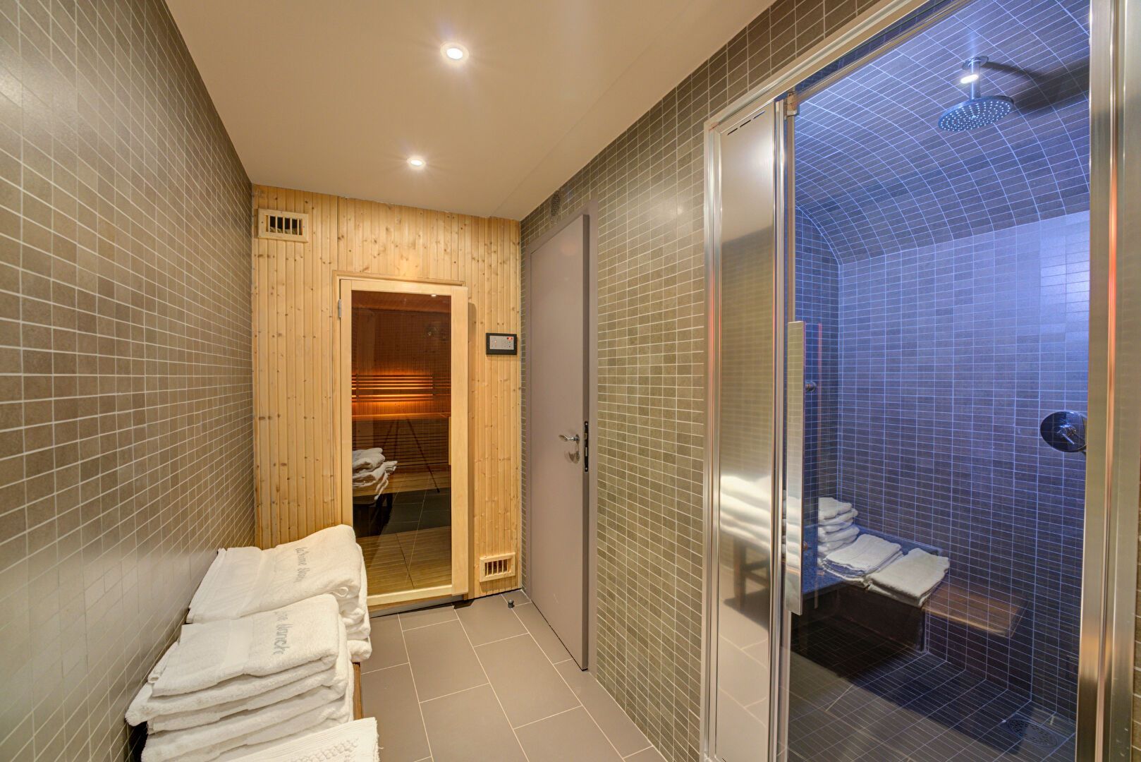 Un couloir de spa avec sauna, douche et serviettes. Sauna en bois clair, douche à éclairage bleu et murs carrelés gris.