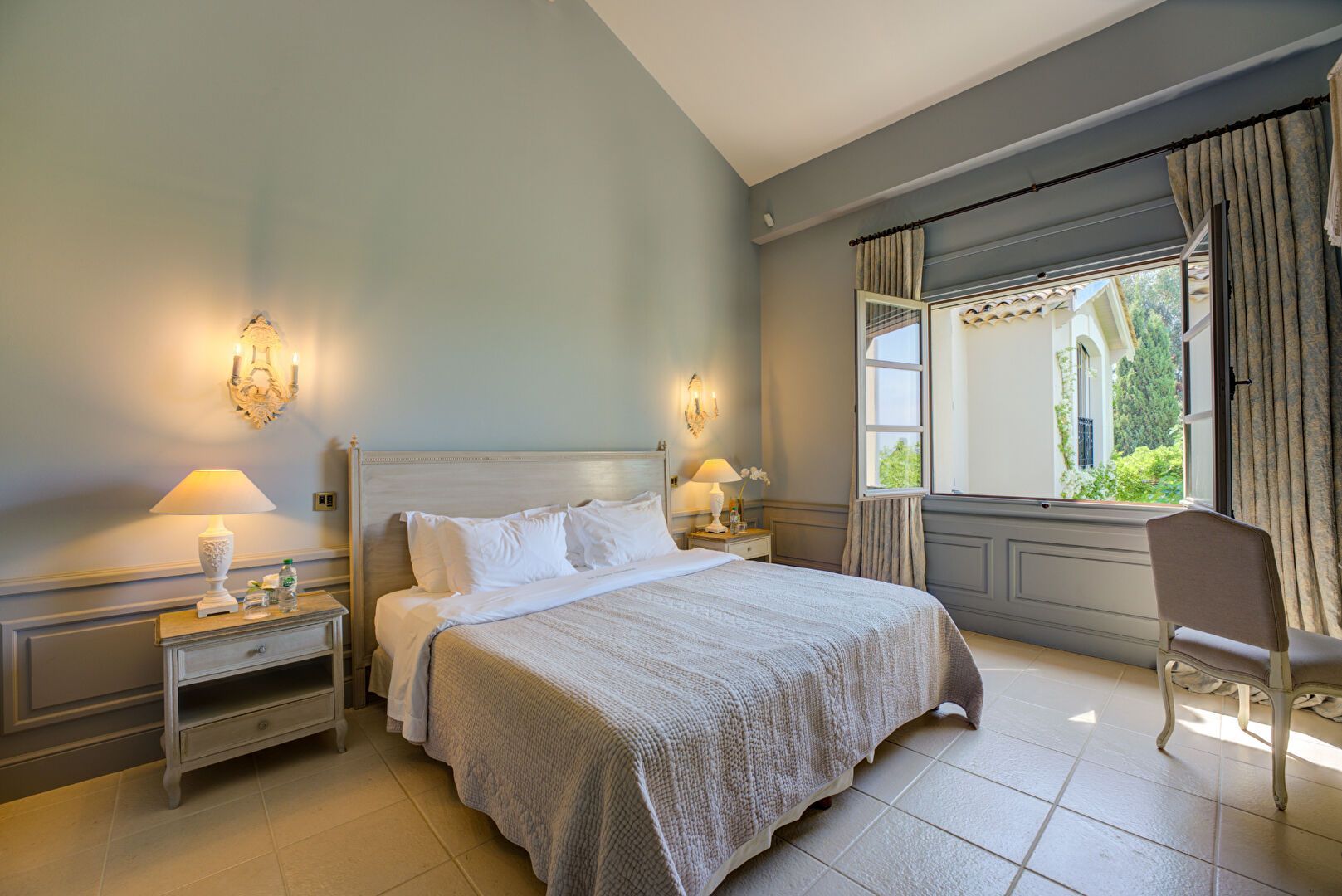 Chambre avec un lit, des tables de chevet et une fenêtre avec vue. Murs gris clair et mobilier de couleur claire.