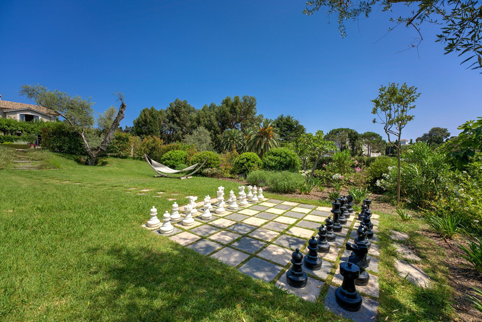 Un jeu d'échecs géant se dresse dans un jardin ensoleillé, avec des pièces noires et blanches sur un plateau carré. De l'herbe verte et des arbres entourent le tout.