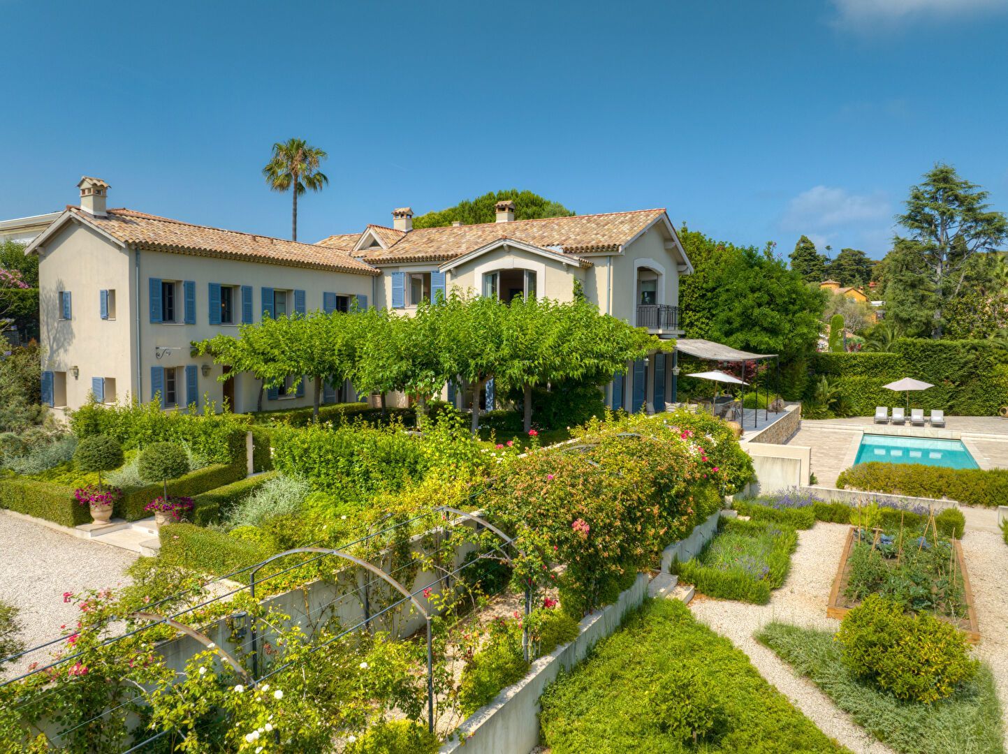 Maison de style méditerranéen avec volets bleus, piscine et jardins soignés sous un ciel d'un bleu limpide.
