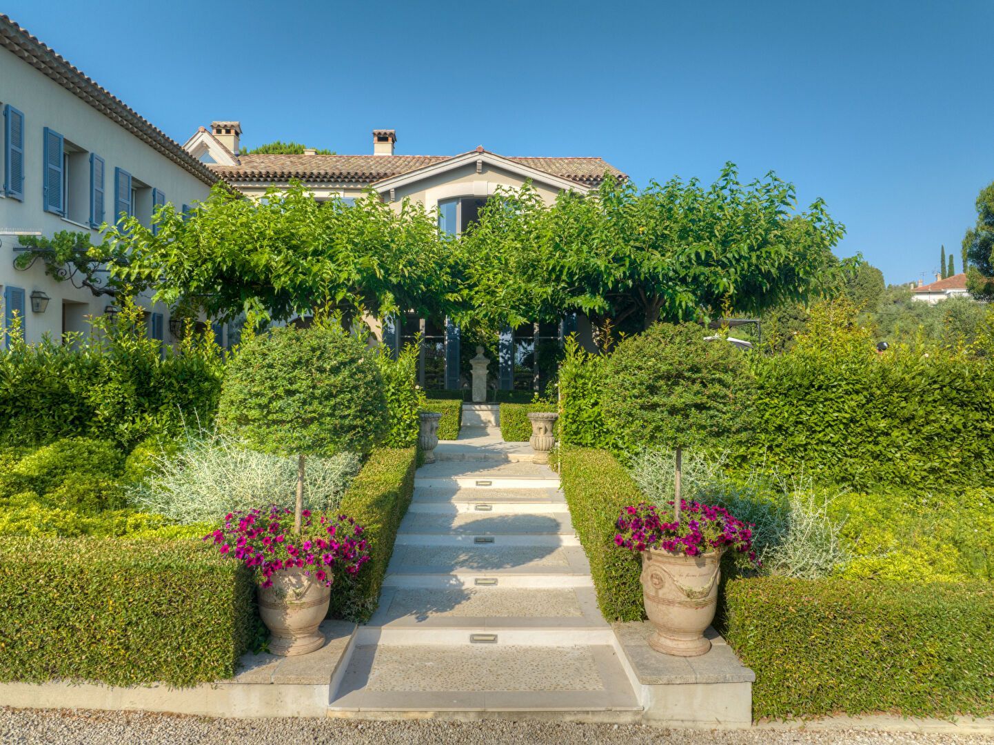 Un escalier en pierre mène à une maison en traversant un jardin soigné avec des fleurs en pot, des haies taillées et une végétation luxuriante.