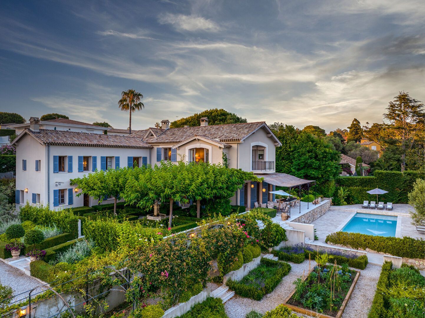 Élégante maison à deux étages avec volets bleus, piscine et parterres de fleurs sous un ciel de coucher de soleil.