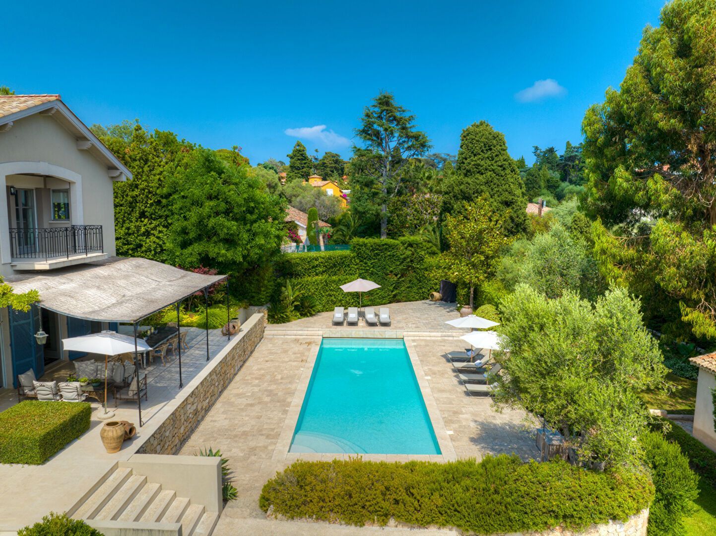 Une piscine rectangulaire entourée d'une terrasse en pierre et de chaises longues. Une villa se trouve à gauche, entourée d'une végétation luxuriante.