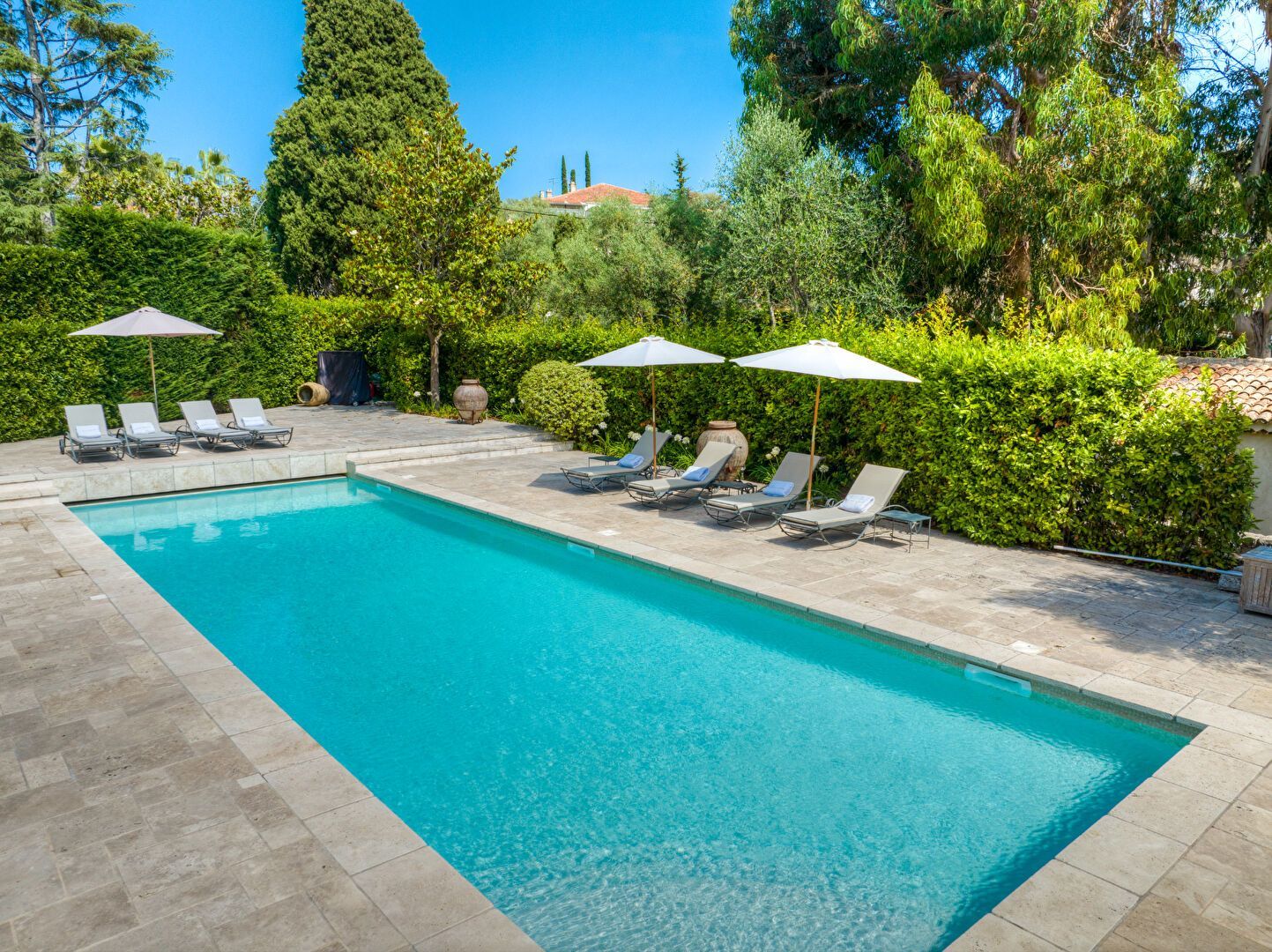 Piscine rectangulaire entourée de chaises longues, de parasols et d'une végétation luxuriante par une journée ensoleillée.