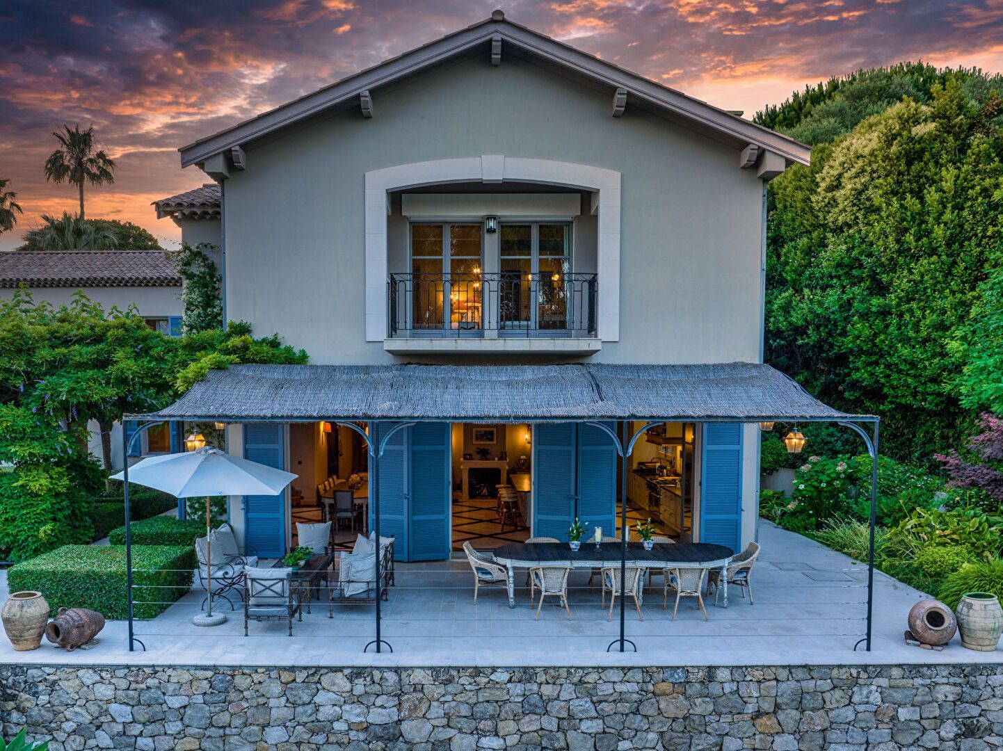Maison à deux étages avec volets bleus, patio et table à manger, sur fond de ciel de coucher de soleil.