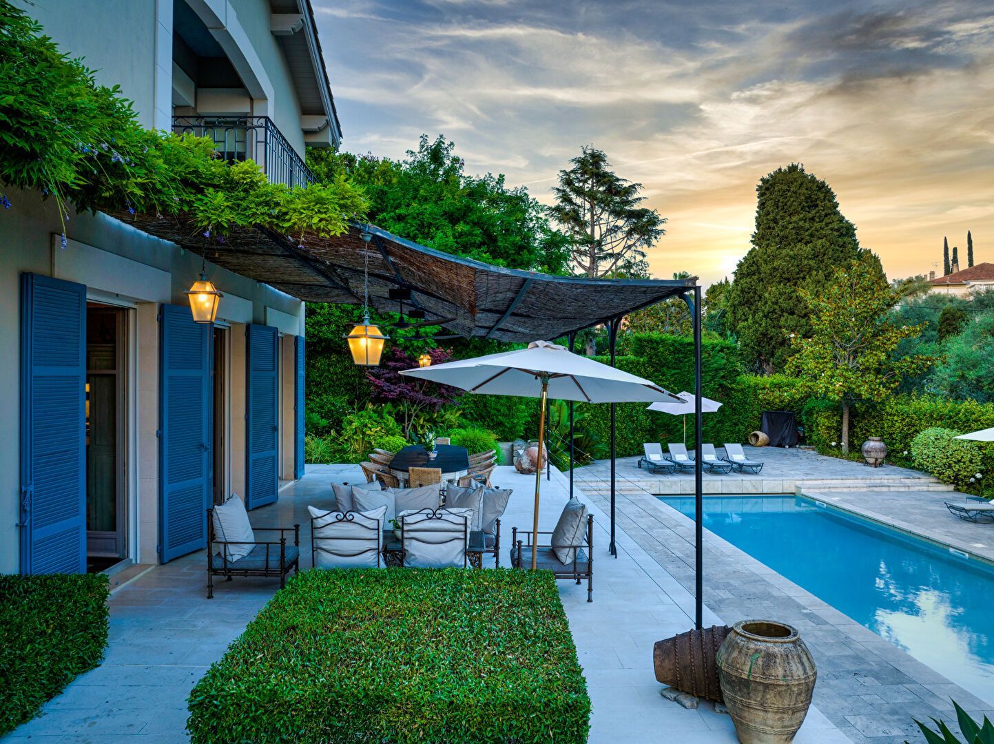 Patio avec sièges, piscine et verdure dans une villa aux volets bleus, sous un ciel de coucher de soleil.
