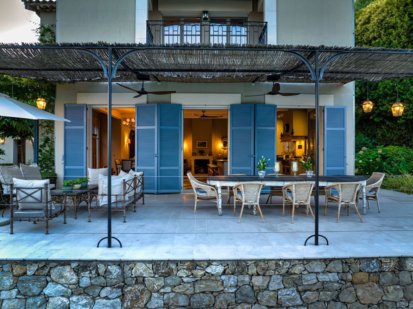 Patio d'une maison avec des volets bleus, une longue table et un mur en pierre.