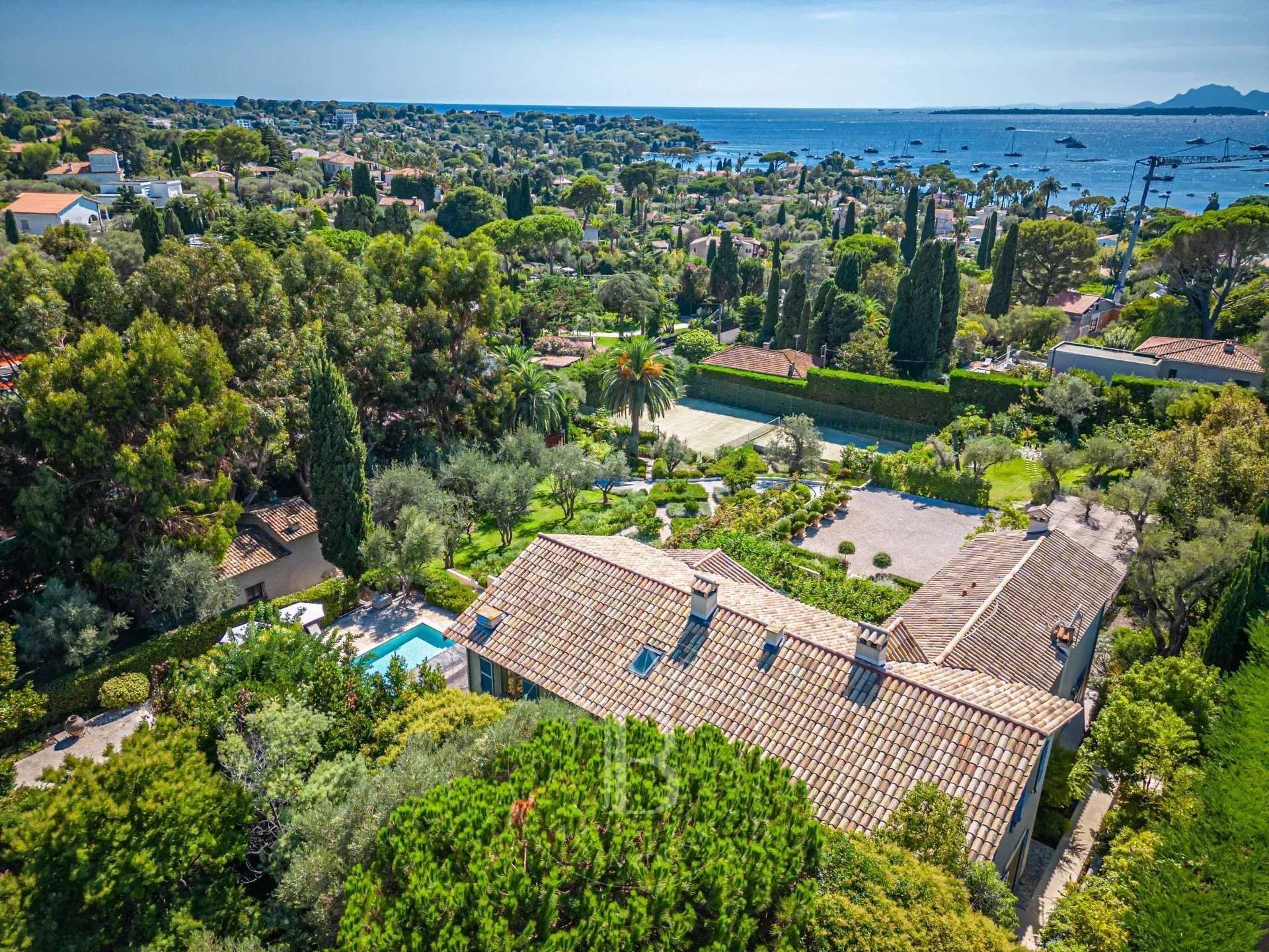 Vue aérienne d'une villa avec toit de tuiles, piscine, court de tennis et végétation luxuriante donnant sur la mer.