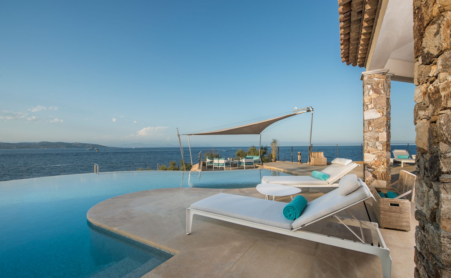 Piscine à débordement avec vue sur l'océan ; chaises longues, table ombragée et murs en pierre.