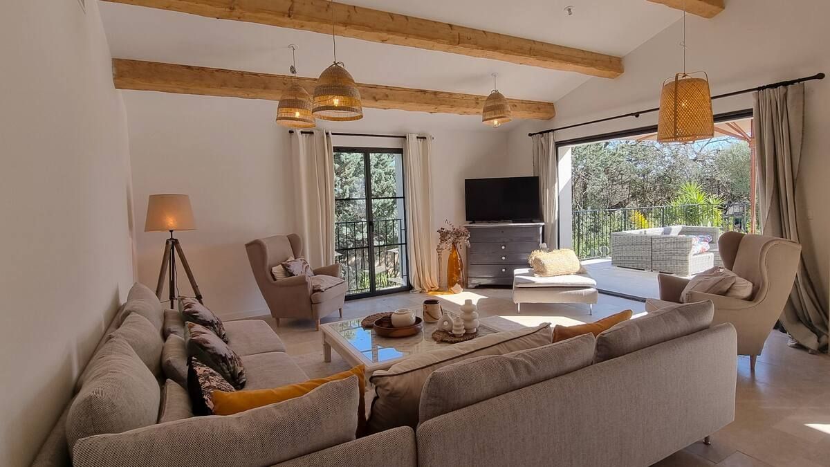 Salon avec un grand canapé d'angle gris, une table basse en verre et des fenêtres ouvertes donnant sur la verdure. Murs beiges, poutres apparentes et suspensions lumineuses.