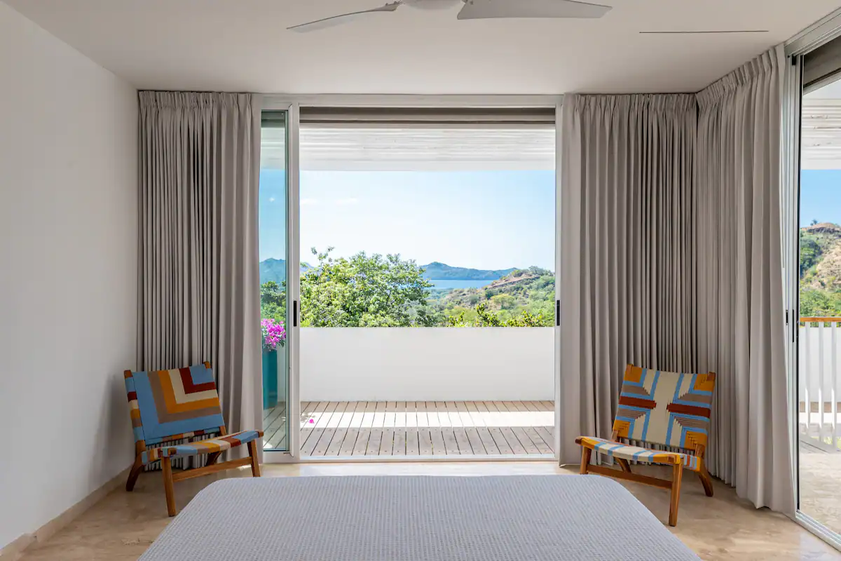 Chambre avec portes vitrées ouvertes donnant sur un balcon offrant une vue panoramique ; deux chaises colorées.