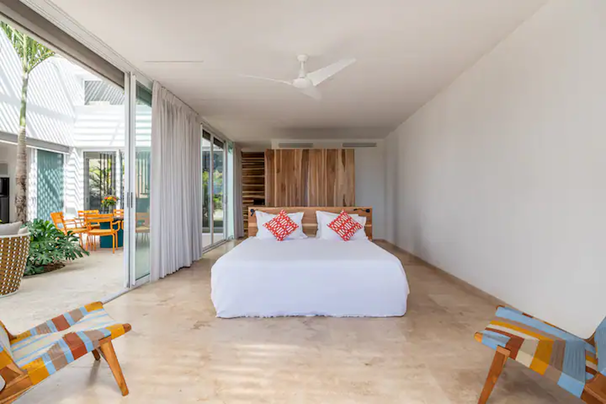 Chambre avec un lit, une tête de lit en bois, deux chaises à motifs et des portes coulissantes ouvertes donnant sur une cour.