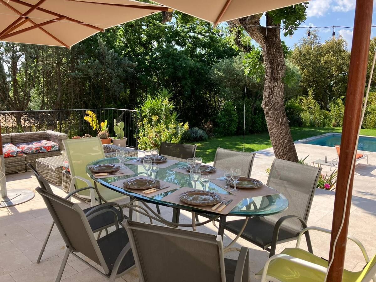 Table à manger extérieure dressée pour un repas, sous un grand parasol sur une terrasse donnant sur une piscine et une végétation luxuriante.