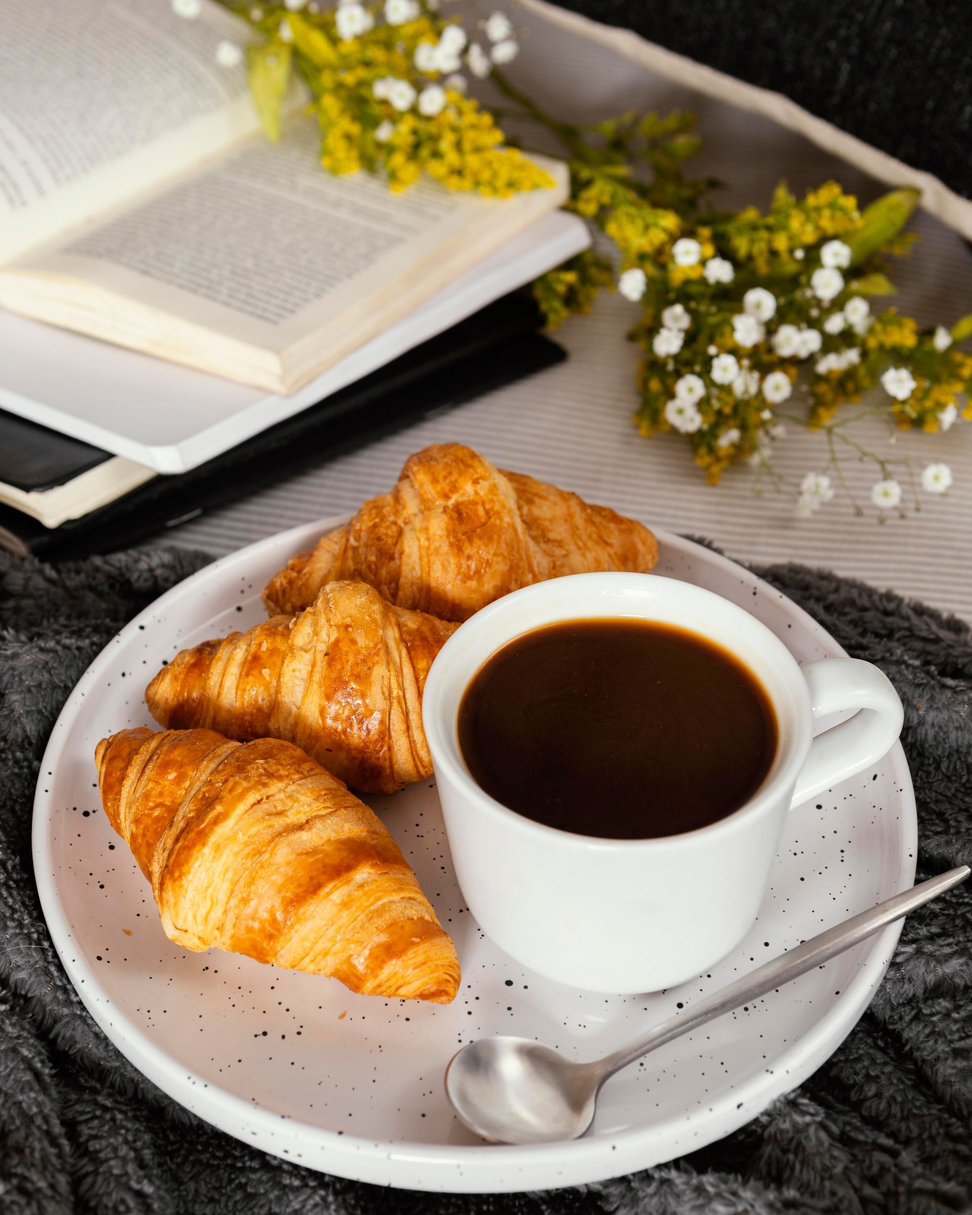 Croissants, café et un livre posé sur une assiette fleurie : une scène de petit-déjeuner chaleureuse.