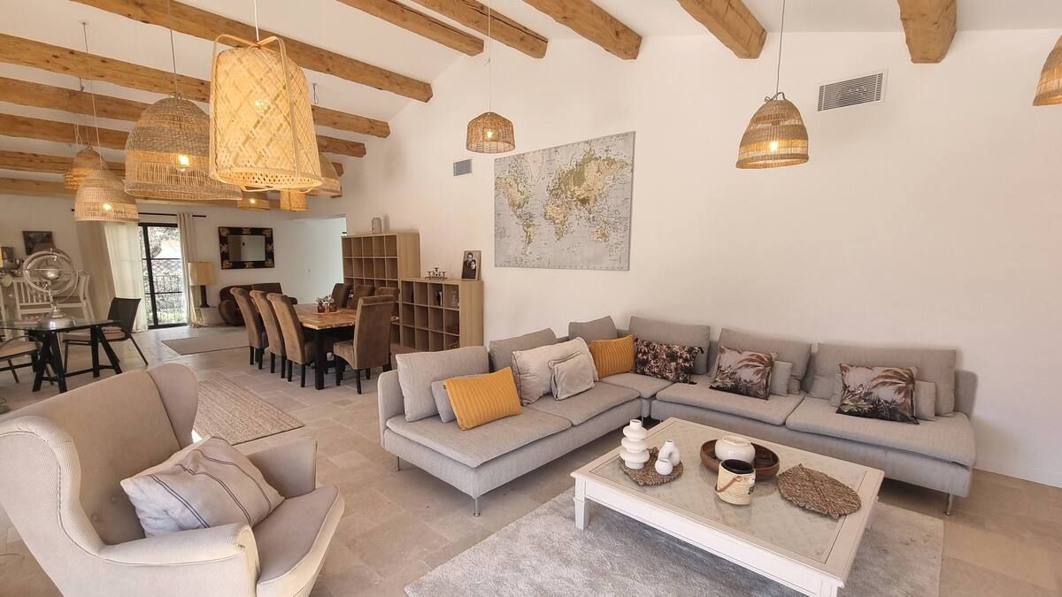 Salon avec plafond voûté, poutres apparentes et lampes en osier. Canapé beige, fauteuil et table à manger.