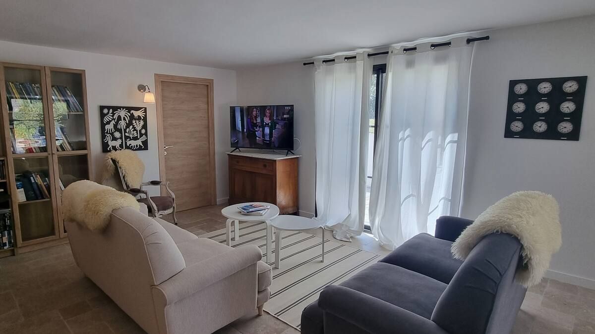 Salon avec deux canapés, télévision, tapis et grandes fenêtres avec rideaux blancs.