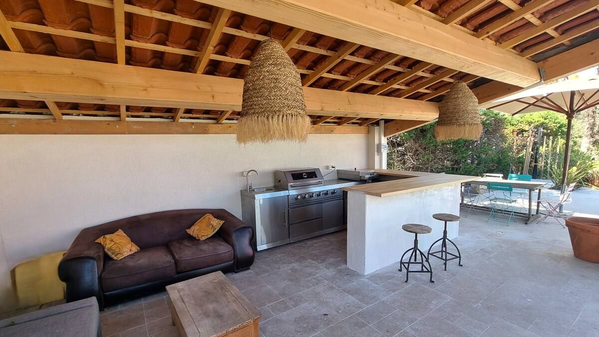 Cuisine extérieure avec barbecue intégré, bar, sièges et luminaires suspendus en osier sous une pergola en bois.