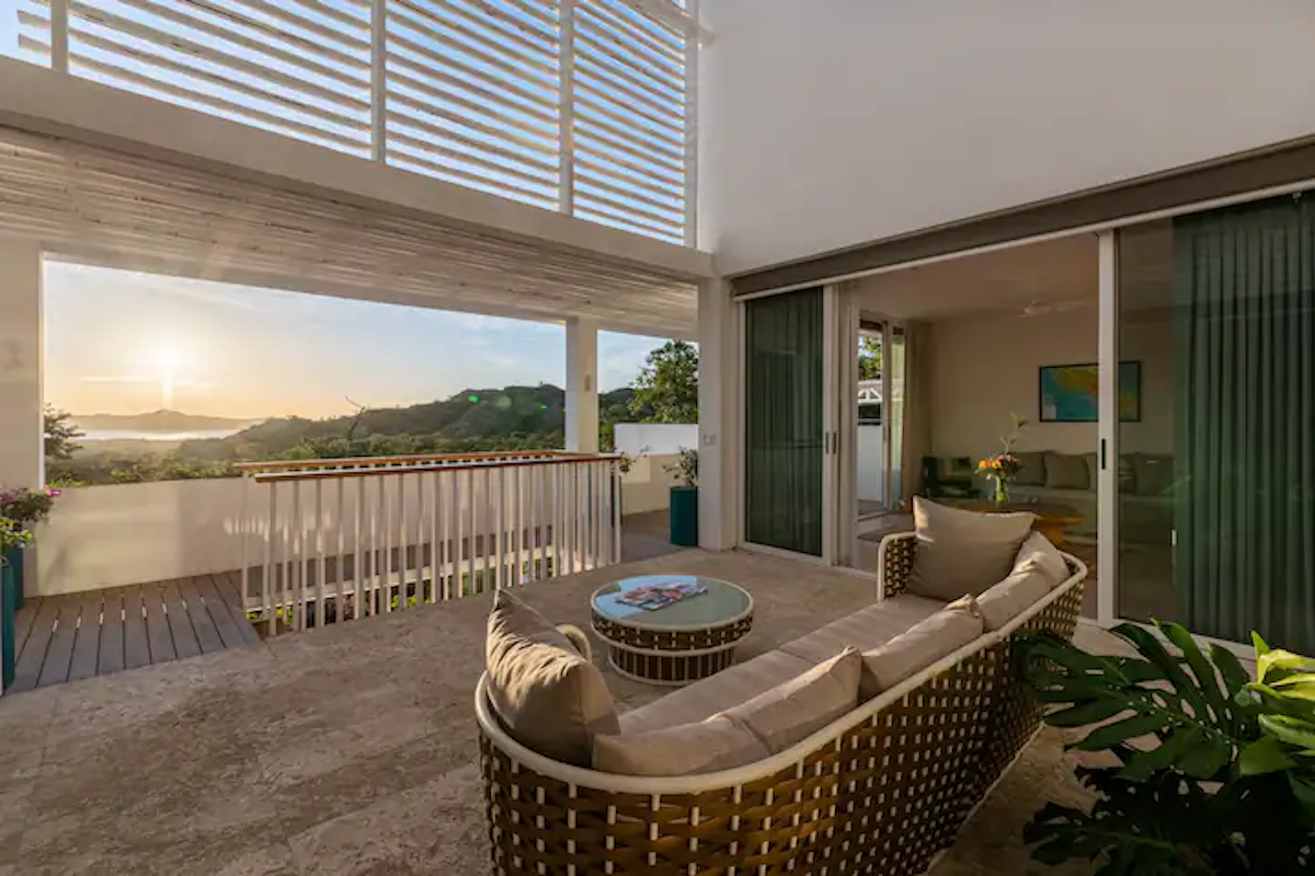 Terrasse extérieure avec canapé en osier, petite table, donnant sur un paysage pittoresque. Des portes coulissantes mènent à l'intérieur.